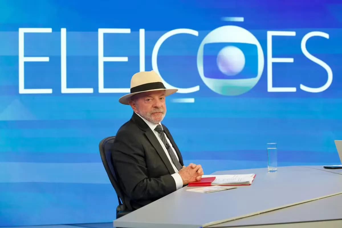 O presidente Lula, de chapéu, sentado à bancada do Jornal Nacional diante do painel "Eleições"